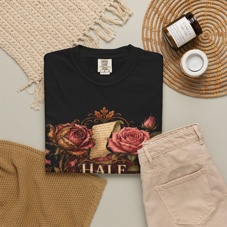 A folded black graphic T-shirt is arranged in a styled flat lay with beige pants, a woven blanket, a candle jar, and a small cup on a light surface. The shirt graphic features pink roses, a parchment scroll, and the partial text &