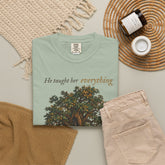 A folded sage green graphic T-shirt is styled in a flat lay with beige pants, a woven blanket, a candle jar, and a small cup on a neutral surface. The shirt features the text &