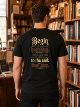 A man viewed from behind wearing a black graphic t-shirt in a cozy, book-filled library or bookstore. The back of the shirt features a retro-style typographic design with the words &