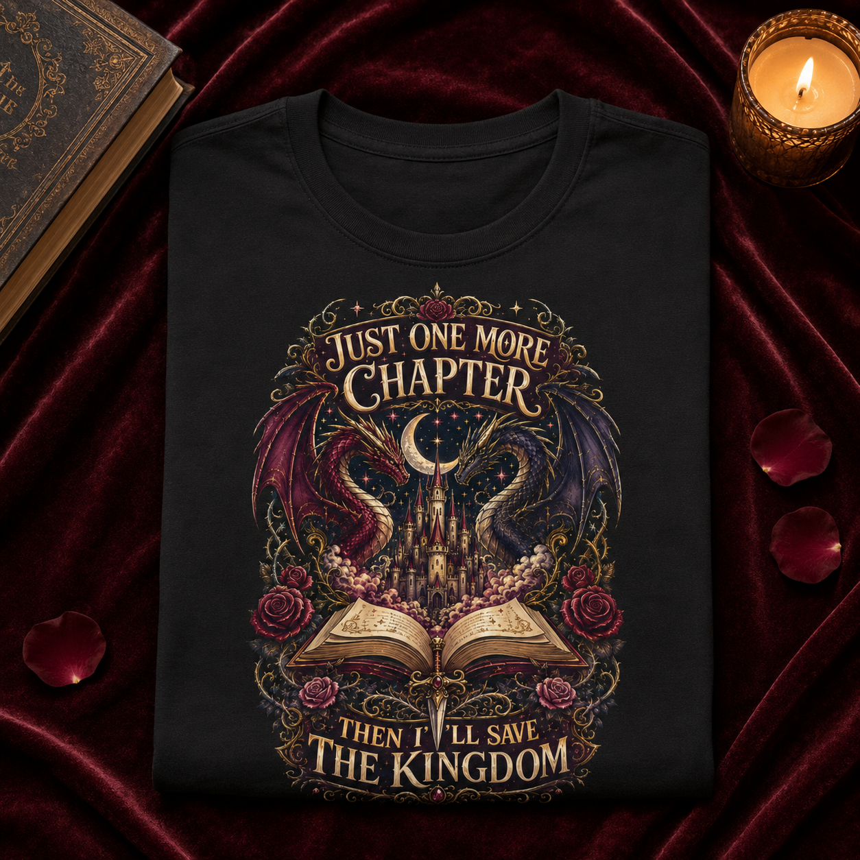A black short-sleeve graphic T-shirt laid flat on a dark red velvet surface, surrounded by decorative props including an old book, a lit candle, and scattered rose petals. The shirt features a large fantasy-style print with a castle, open book, roses, and the text &