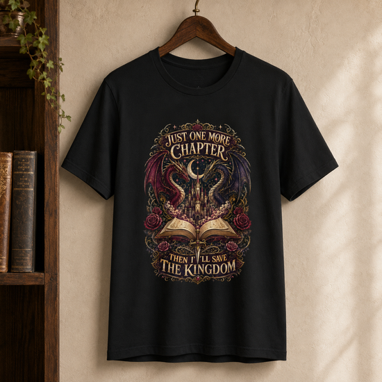 A black short-sleeve graphic T-shirt hanging on a wooden hanger against a textured beige wall, with a bookshelf visible on the left. The shirt features a large fantasy-style front print with a dragon, open book, roses, and the text &