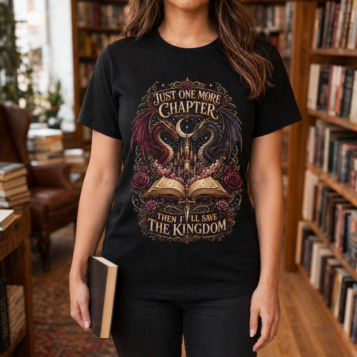 A woman wearing a black graphic t-shirt in a cozy, book-filled library setting. The t-shirt features a detailed fantasy illustration of a witch&
