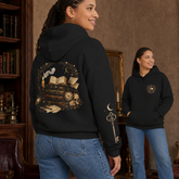 A woman wearing a black hoodie with a gothic-themed back graphic featuring an open book, skull, and key, shown in a warm indoor setting with a mirror reflecting her front view of the hoodie.