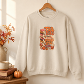 An off-white crewneck sweatshirt hanging on a wooden hanger against a warm beige wall. The sweatshirt features a colorful graphic on the front with a stack of books, pumpkins, autumn leaves, and a coffee cup, with the text &