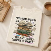 A folded cream-colored t-shirt with a humorous book-themed graphic and quote, styled in a cozy flat-lay setting with a book, a cup of coffee, and a knitted blanket.