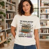 A woman wearing a white graphic t-shirt with a library-themed social media quote and book illustration, standing in a book-filled room.
