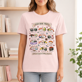 A woman wearing a light pink graphic t-shirt with a colorful cartoon illustration of sushi tropes and cat characters. The background shows a softly blurred indoor setting with bookshelves and a plant.