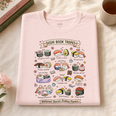 A light pink folded t-shirt with a colorful cartoon sushi-themed graphic print, styled on a cream fabric background with books and a cup in the corners.