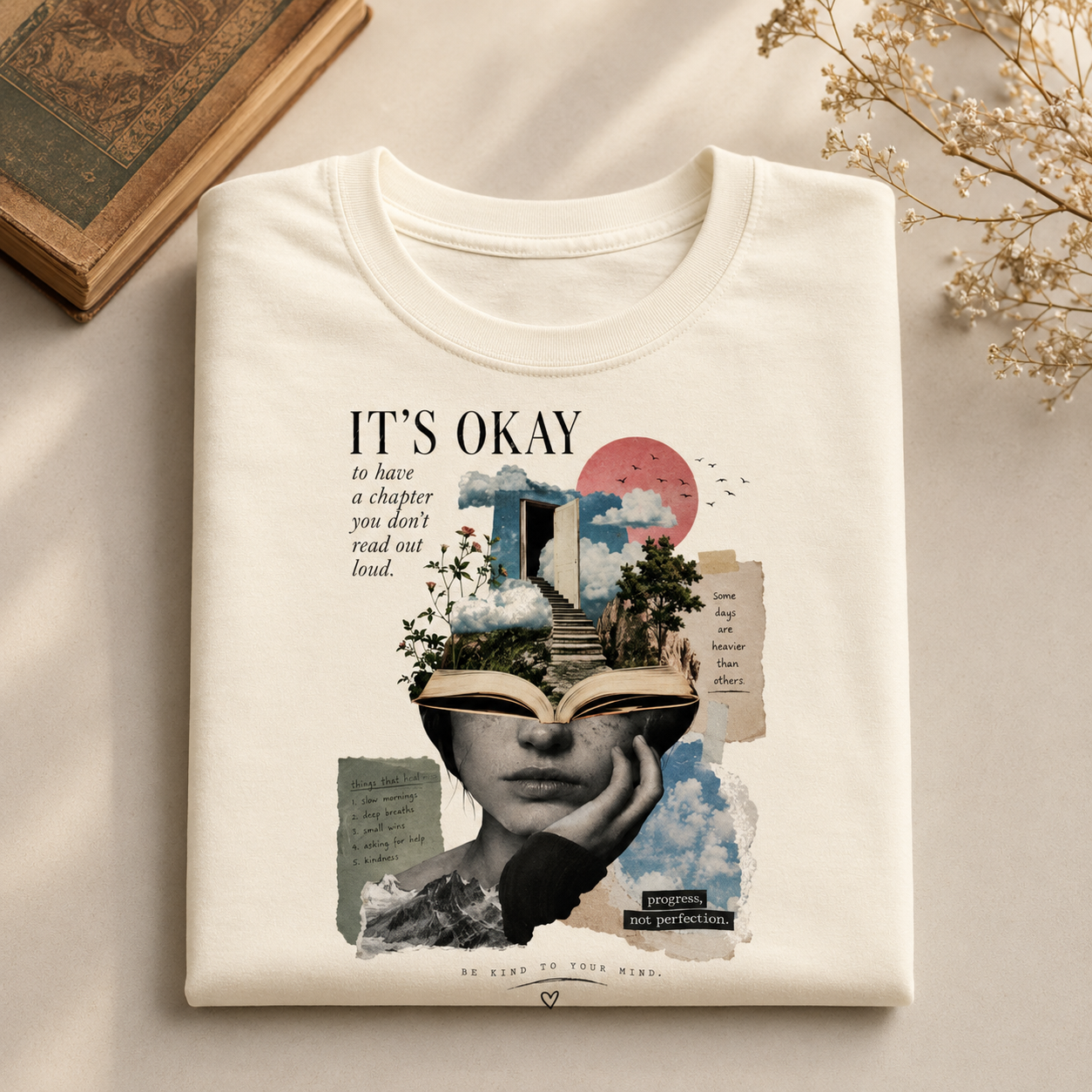 A folded off-white graphic T-shirt is displayed on a light beige surface with styled props, including an old book and dried flowers. The shirt features a detailed collage graphic with the text &