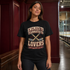 A woman wearing a black graphic t-shirt with a hockey-themed slogan, standing in a hallway with dark red wood paneling and an indoor sports arena visible in the background.