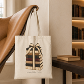 A cream canvas tote bag with a graphic of a stack of books tied with a ribbon, hanging from the arm of a brown leather chair in a cozy, softly lit room with a bookshelf in the background.