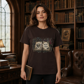 A woman stands in a cozy, dimly lit library wearing a dark brown graphic t-shirt with a vintage-style open book design on the front. The background features wooden bookshelves filled with books, a leather armchair, and a window.