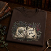 A folded dark brown t-shirt with a gothic-style graphic of an open book surrounded by flowers and birds, featuring the text &