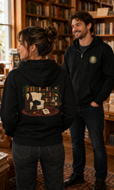 A man and a woman standing in a cozy, book-filled library. The woman in the foreground is shown from the back wearing a black hoodie with a large, detailed graphic of a library scene on the back.