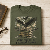 An olive green t-shirt with a detailed graphic print of an eagle over books, swords, and money, laid flat on a light wood background next to a book and glasses.