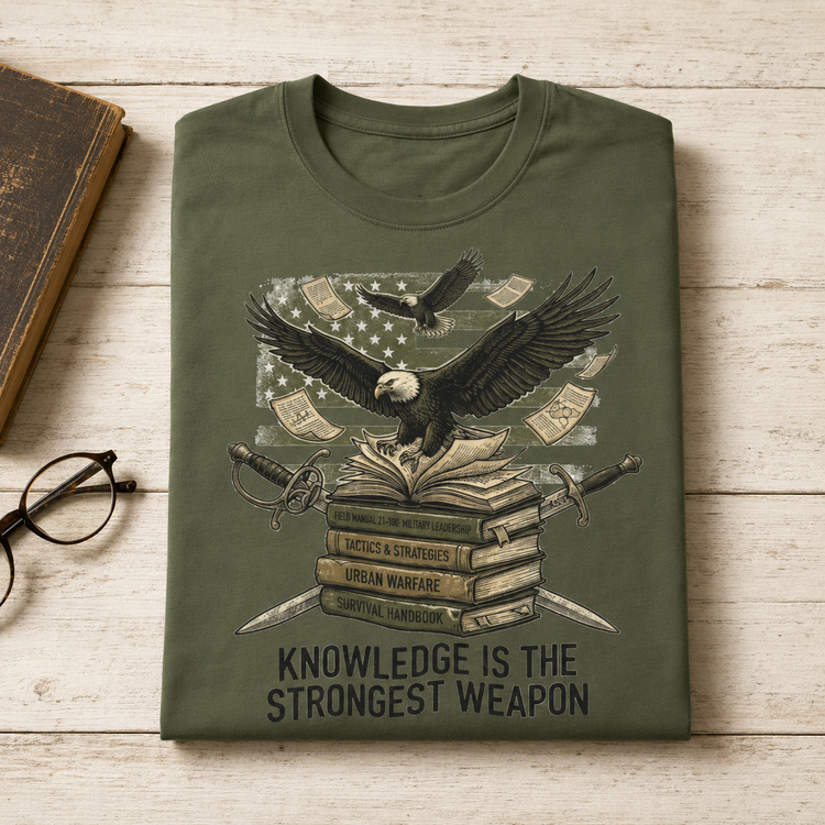 An olive green t-shirt with a detailed graphic print of an eagle over books, swords, and money, laid flat on a light wood background next to a book and glasses.