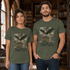 A man and a woman wearing matching olive green graphic t-shirts in a library setting. The shirts feature a graphic of a raven perched on a stack of books with a skull and American flag, and the text &