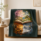 A dark teal throw blanket draped over a beige sofa in a cozy living room setting. The blanket features a large, colorful printed design of an open book with four distinct seasons: pink cherry blossoms, a sunny landscape, autumn leaves, and a snowy winter scene.