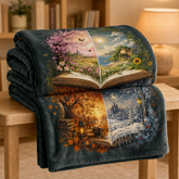 A folded dark blanket draped over a wooden table in a cozy indoor setting. The blanket features a large printed design of an open book with four illustrated scenes: a pink cherry blossom landscape, a blue lake with a house, a warm autumn campfire scene, and a snowy winter forest.