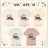 A color selection graphic for a graphic t-shirt showing five different color options: Natural, Heather Dust, Soft Cream, Heather Prism Mint, and Heather Mauve. Each option displays a short-sleeve crewneck t-shirt with a book stack and coffee cup graphic and the text &