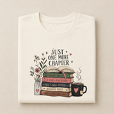 A folded off-white crewneck t-shirt with a graphic print on the front. The print features an illustration of an open book on a stack of four colorful books next to a mason jar with wildflowers and a black coffee mug with a pink heart.