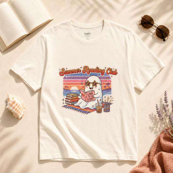 An off-white short-sleeve graphic T-shirt laid flat on a beige surface with styled props including sunglasses, an open book, a seashell, dried lavender, and a pink knit fabric. The shirt has a colorful retro-style reading club illustration with the text &