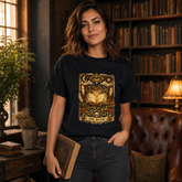 A woman stands in a cozy, dimly lit library wearing a black graphic t-shirt with a gold and brown illustration of an open book under an eye motif, holding a vintage book. The background features bookshelves and a leather armchair.