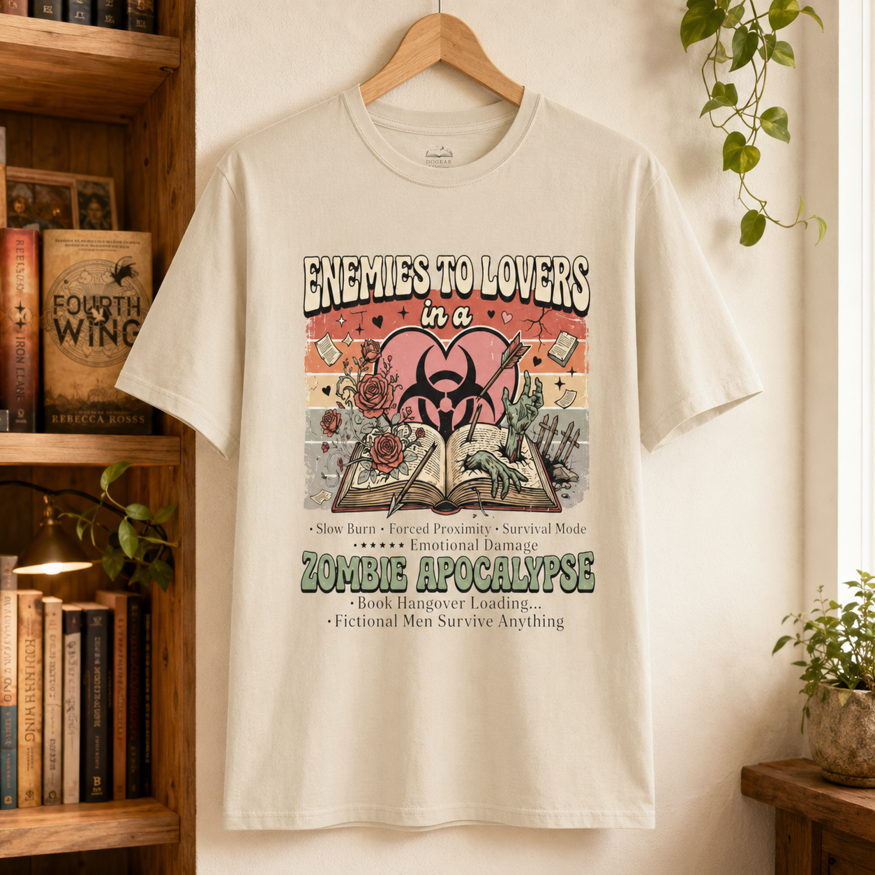 A cream-colored graphic t-shirt hanging on a wooden hanger against a warm wall, with a bookshelf and houseplants visible in the background. The t-shirt features a detailed graphic with the text &