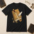 A black short-sleeve t-shirt laid flat on a textured cream background, featuring a graphic print of an antique gold book, a silver dagger, and a white feather quill. The shirt is styled with vintage props including books, glasses, and dried leaves.