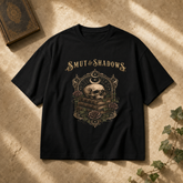 A black short-sleeve graphic T-shirt laid flat on a textured beige surface with warm sunlight shadows. The shirt features a gothic-style print with the words &