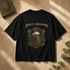 A black short-sleeve graphic T-shirt laid flat on a textured beige surface with warm sunlight shadows. The shirt features a gothic-style print with the words &