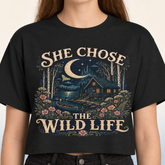 A woman wearing a black cropped t-shirt with a detailed graphic print. The graphic features a rustic cabin by a lake under a crescent moon, surrounded by flowers and trees, with the text &
