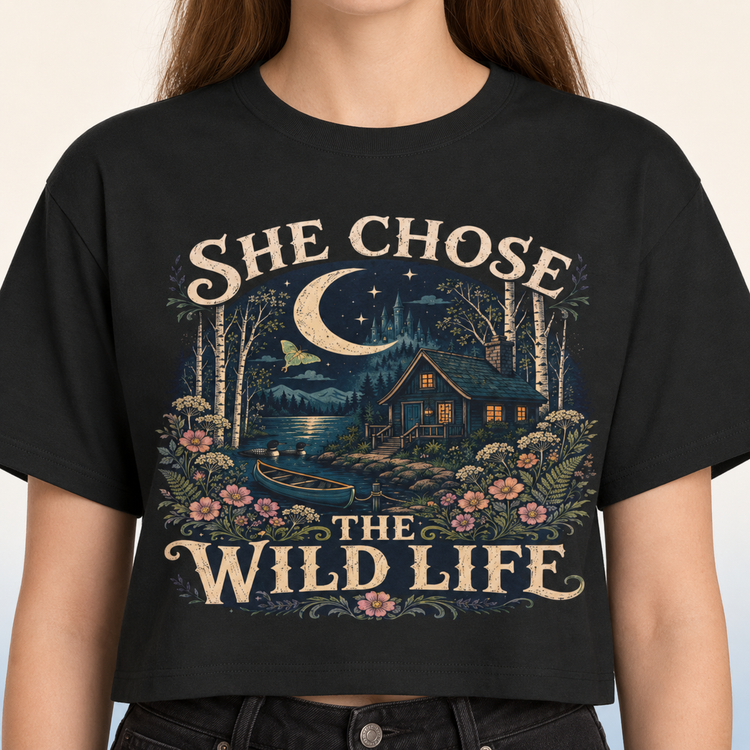 A woman wearing a black cropped t-shirt with a detailed graphic print. The graphic features a rustic cabin by a lake under a crescent moon, surrounded by flowers and trees, with the text &
