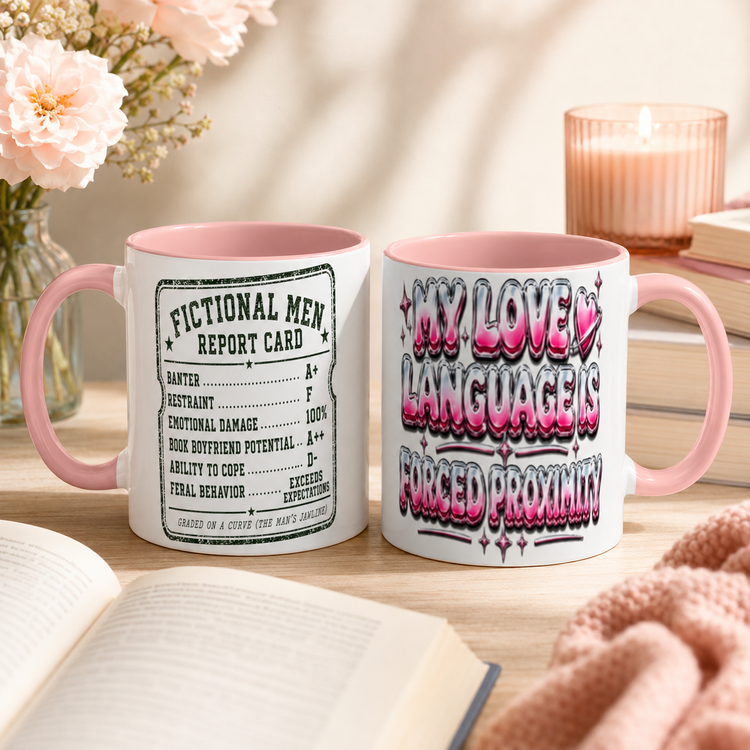 Two ceramic mugs with pink interiors and handles displayed on a wooden table in a cozy styled setting. One mug shows a vintage-style fictional men report card graphic, and the other features bold pink and black graffiti-style text reading &