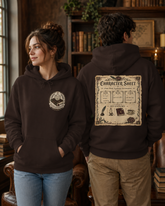 A man and a woman wearing matching dark brown hoodies in a cozy, book-filled library setting. The woman wears a hoodie with a small circular crest on the left chest, while the man wears a hoodie showing a large vintage-style &
