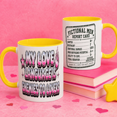 Two white ceramic mugs with yellow interiors and handles are shown on a pink background. The mug in front reads “MY LOVE LANGUAGE IS ENEMIES TO LOVERS” in pink and black bubble lettering, and the mug behind it shows a “Fictional Men Report Card” design.