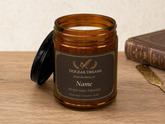 An amber glass jar candle with its black lid leaning beside it on a wooden surface. The front label reads &