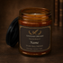 An amber glass jar candle with its black lid leaning beside it on a dark wooden surface. The front label reads &
