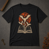 A black short-sleeve t-shirt laid flat on a dark gray textured surface, featuring a graphic print of a vampire bat rising from an open book in front of a red full moon. Decorative props including a vintage key and dried flowers are visible in the corners.