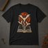 A black short-sleeve t-shirt laid flat on a dark gray textured surface, featuring a graphic print of a vampire bat rising from an open book in front of a red full moon. Decorative props including a vintage key and dried flowers are visible in the corners.