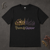 A black short-sleeve graphic t-shirt laid flat on a dark textured background, featuring a split design with a gold floral crown on the left and a purple thorny crown on the right, with the text &