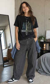 A woman stands in a bedroom setting wearing a black graphic t-shirt with &