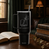 A black insulated travel tumbler with a handle and straw sits on a wooden table in a cozy room setting. The tumbler features a frosted graphic of an open book with crossed swords and the text &