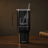 A matte black insulated tumbler with a handle, clear lid, and straw, featuring a white laser-engraved design of an open book and sword with the text &