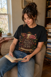A woman sitting in a cozy armchair, reading a book. She is wearing a black graphic t-shirt with a floral design and the text &