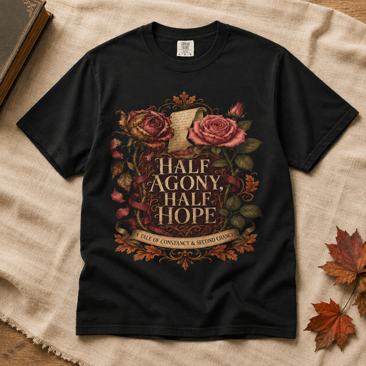 A black short-sleeve graphic T-shirt laid flat on a beige linen surface with autumn leaves and a book corner nearby. The shirt features a large decorative print with roses, leaves, and the text &
