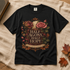A black short-sleeve graphic T-shirt laid flat on a beige linen surface with autumn leaves and a book corner nearby. The shirt features a large decorative print with roses, leaves, and the text &