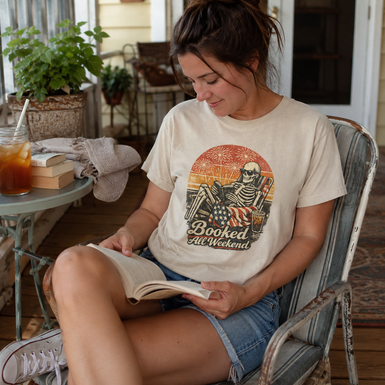 A woman sitting in a rustic chair on a porch, reading a book and wearing a cream-colored graphic t-shirt with a retro skeleton design and the text &