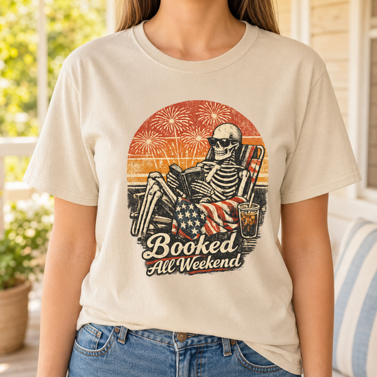 A woman wearing a beige graphic t-shirt with a skeleton lounging in front of a sunset and fireworks design, paired with blue jeans, set against a softly blurred porch background.