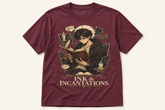 A maroon short-sleeve graphic t-shirt featuring an illustration of a young man reading a book surrounded by ink, quills, and cards, with the text &